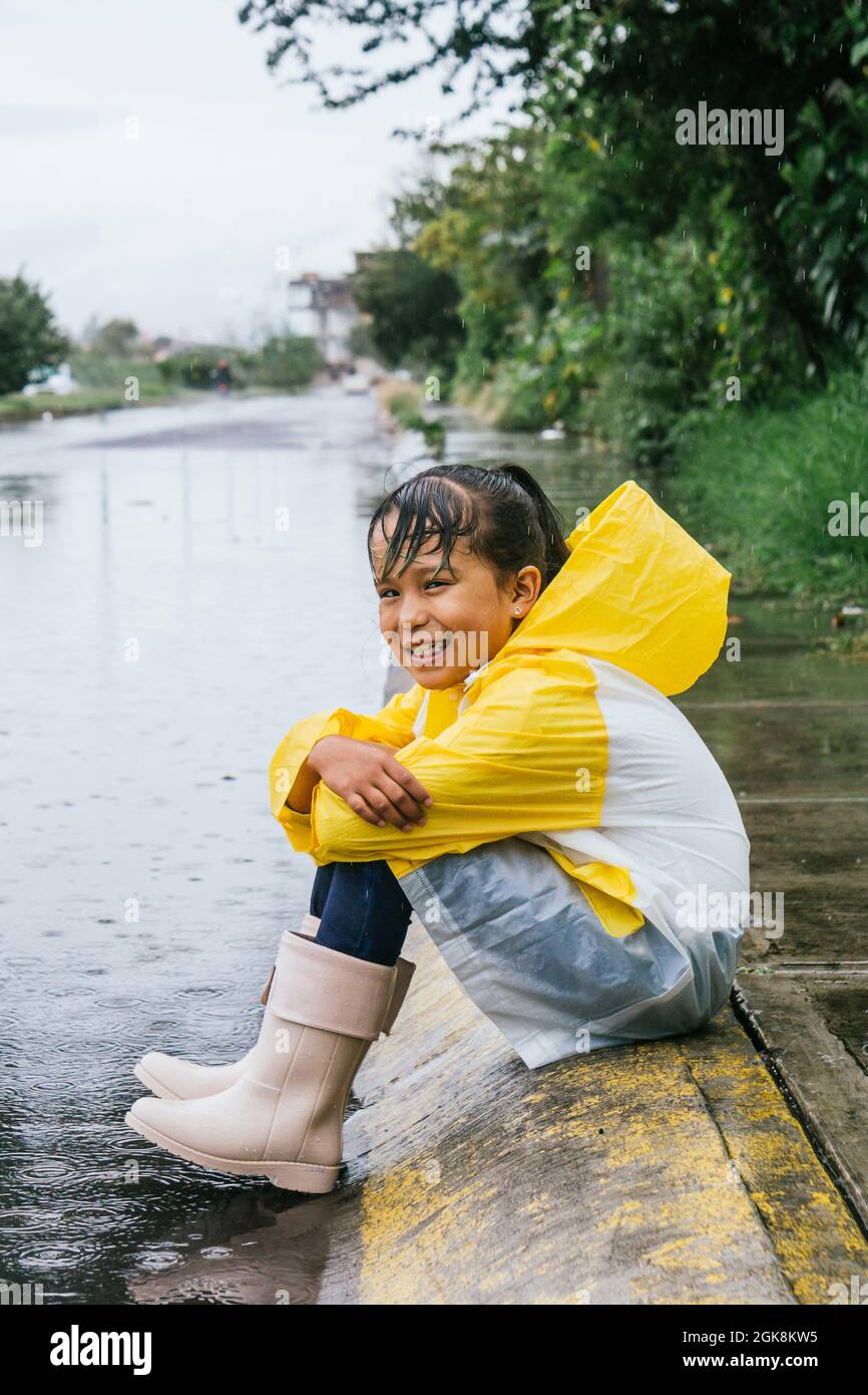 Side view of happy ethnic child in raincoat and gumboots sitting with folded arms on pavement while looking forward on rainy day Stock Photo