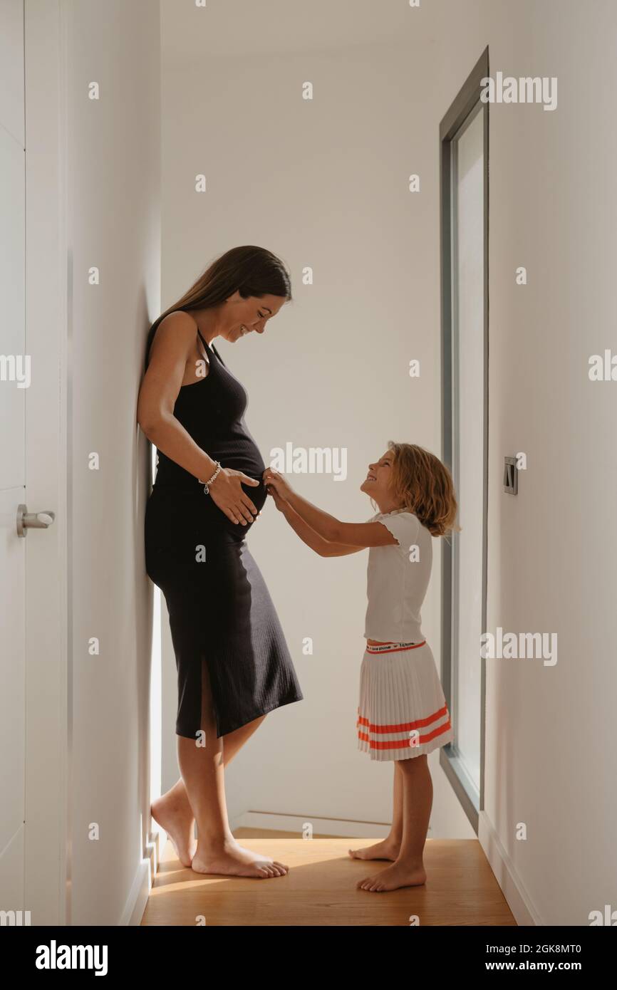 Side view of cheerful barefoot child touching belly of expectant mom while looking at each other in corridor at home Stock Photo