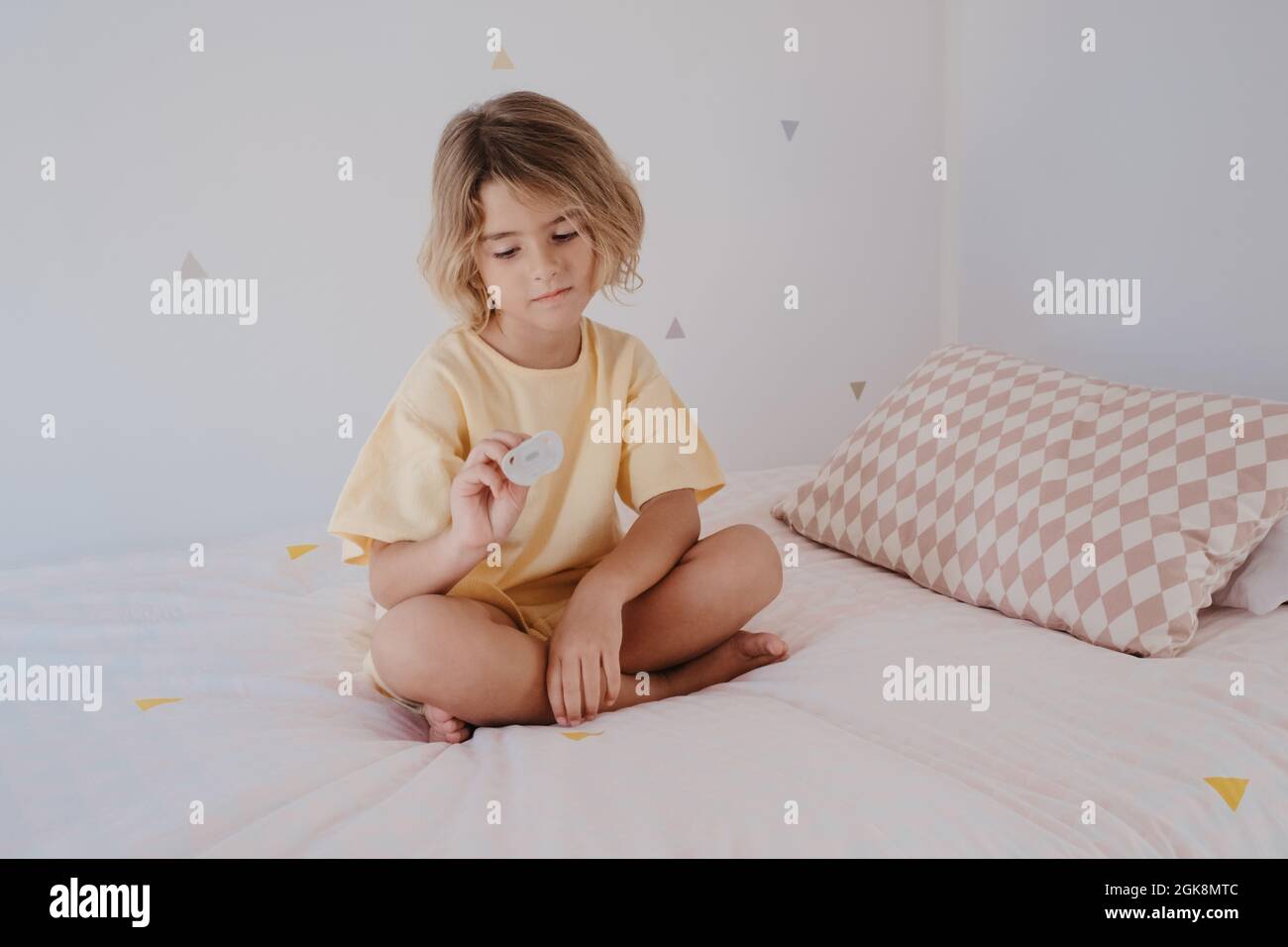 Barefoot child in t shirt with pacifier sitting with crossed legs on soft bed in house Stock Photo