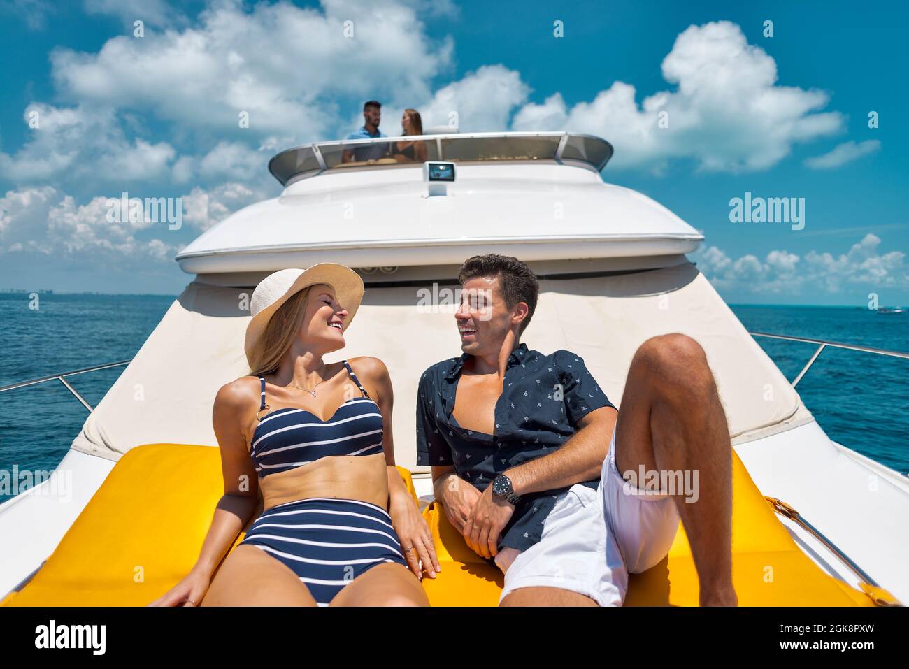 Cheerful young Woman in bikini and hat chatting and flirting with handsome boyfriend while sunbathing together on yacht floating in sea in sunny day d Stock Photo