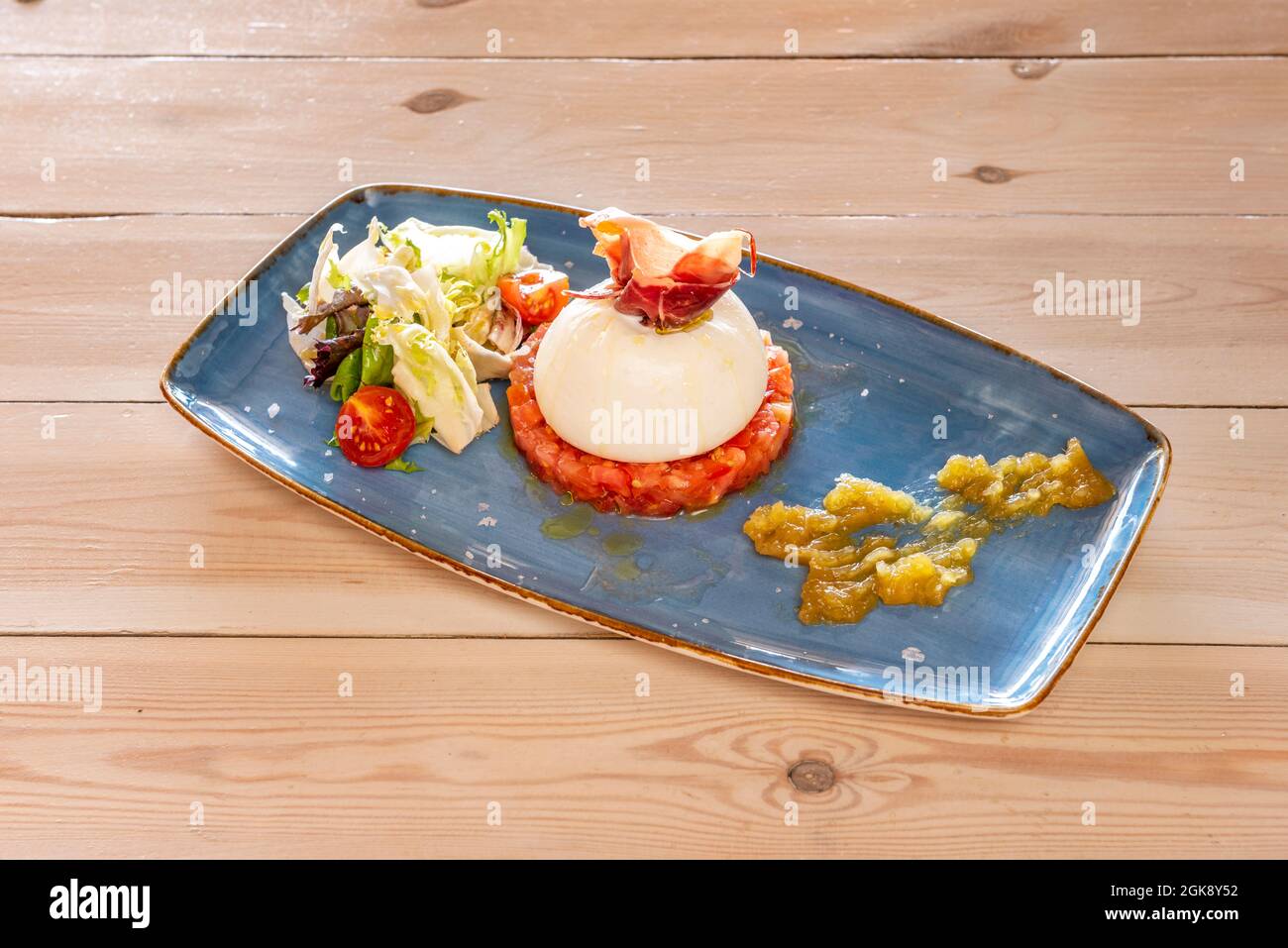 Burrata cheese mounted on tomato tartare, a little Serrano ham and green salad on a blue tray Stock Photo