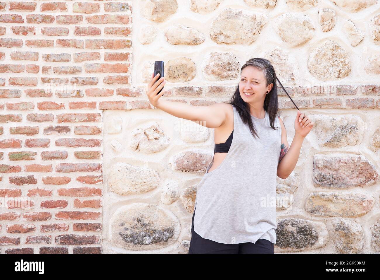 Portrait of a smiling woman taking a selfie with her smartphone outdoors while touching her hair. Stock Photo