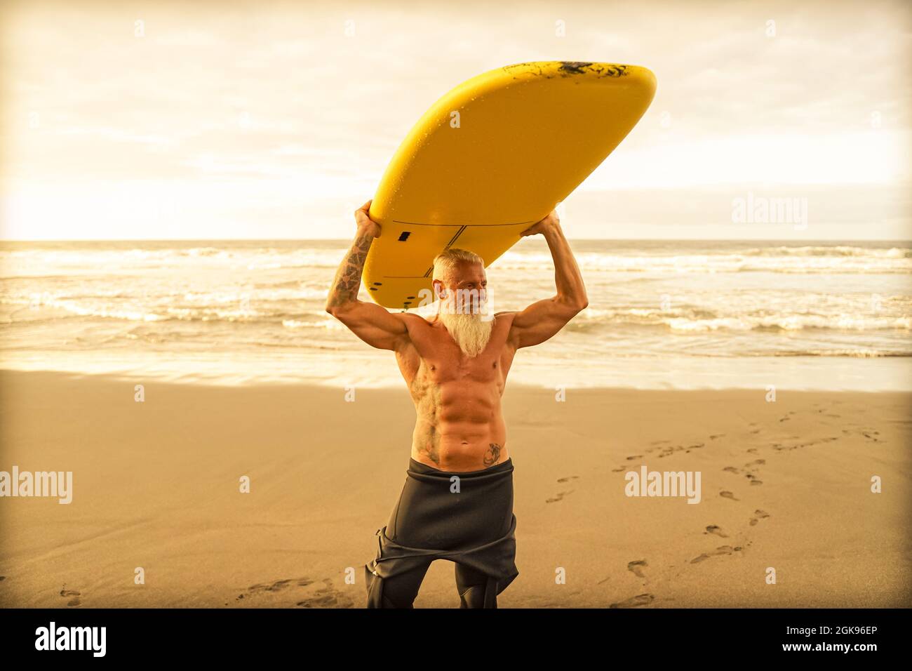 Happy fit senior having fun surfing at sunset time. Sporty bearded man training with surfboard on the beach. Elderly healthy people lifestyle and extr Stock Photo