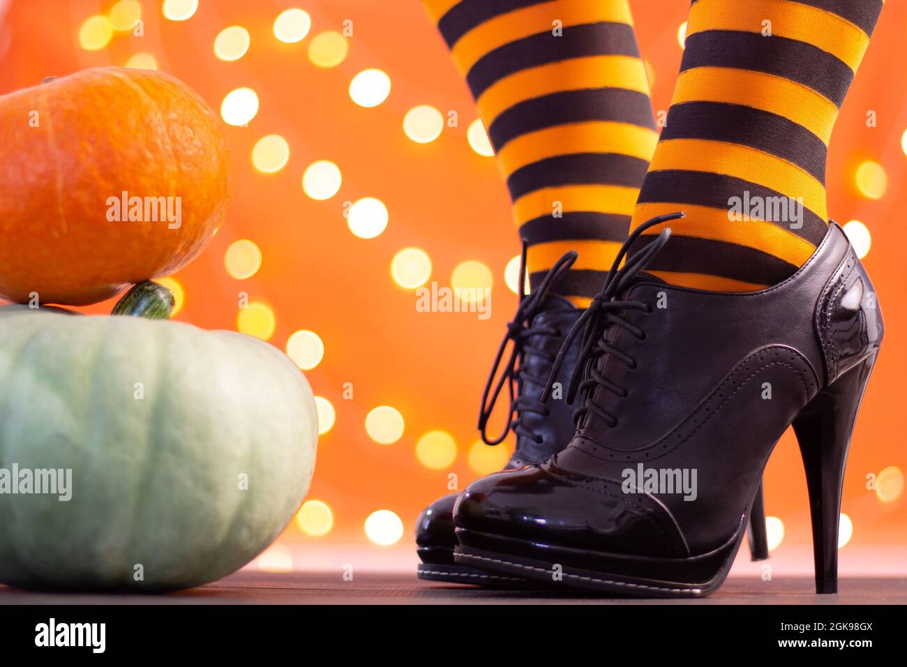 Witch legs in striped stockings and high heel shoes with Halloween pumpkins on an orange background with bokeh. Copy space. Stock Photo