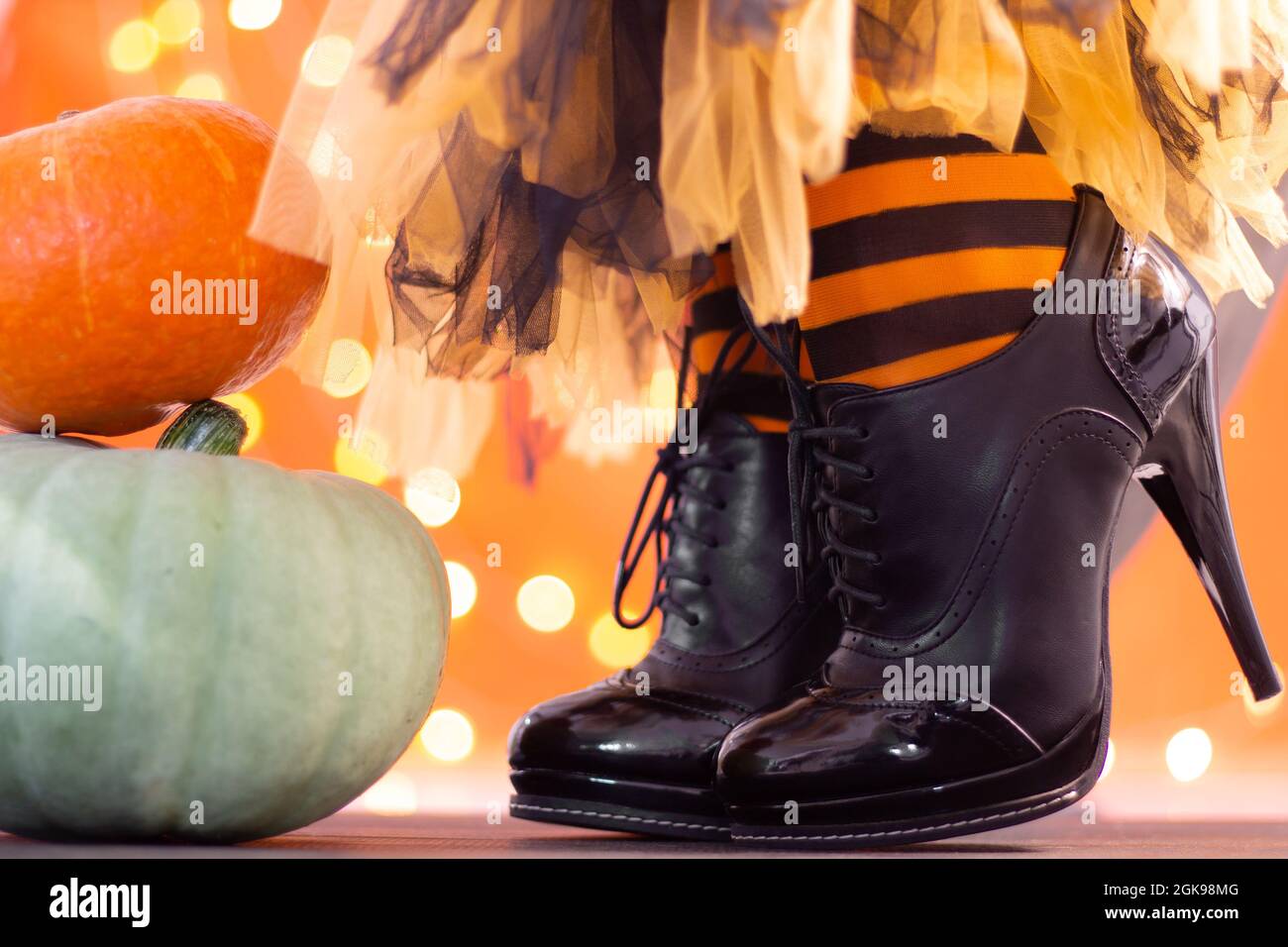 Witch legs in striped stockings and high heel shoes with Halloween pumpkins on an orange background with bokeh. Copy space. Stock Photo