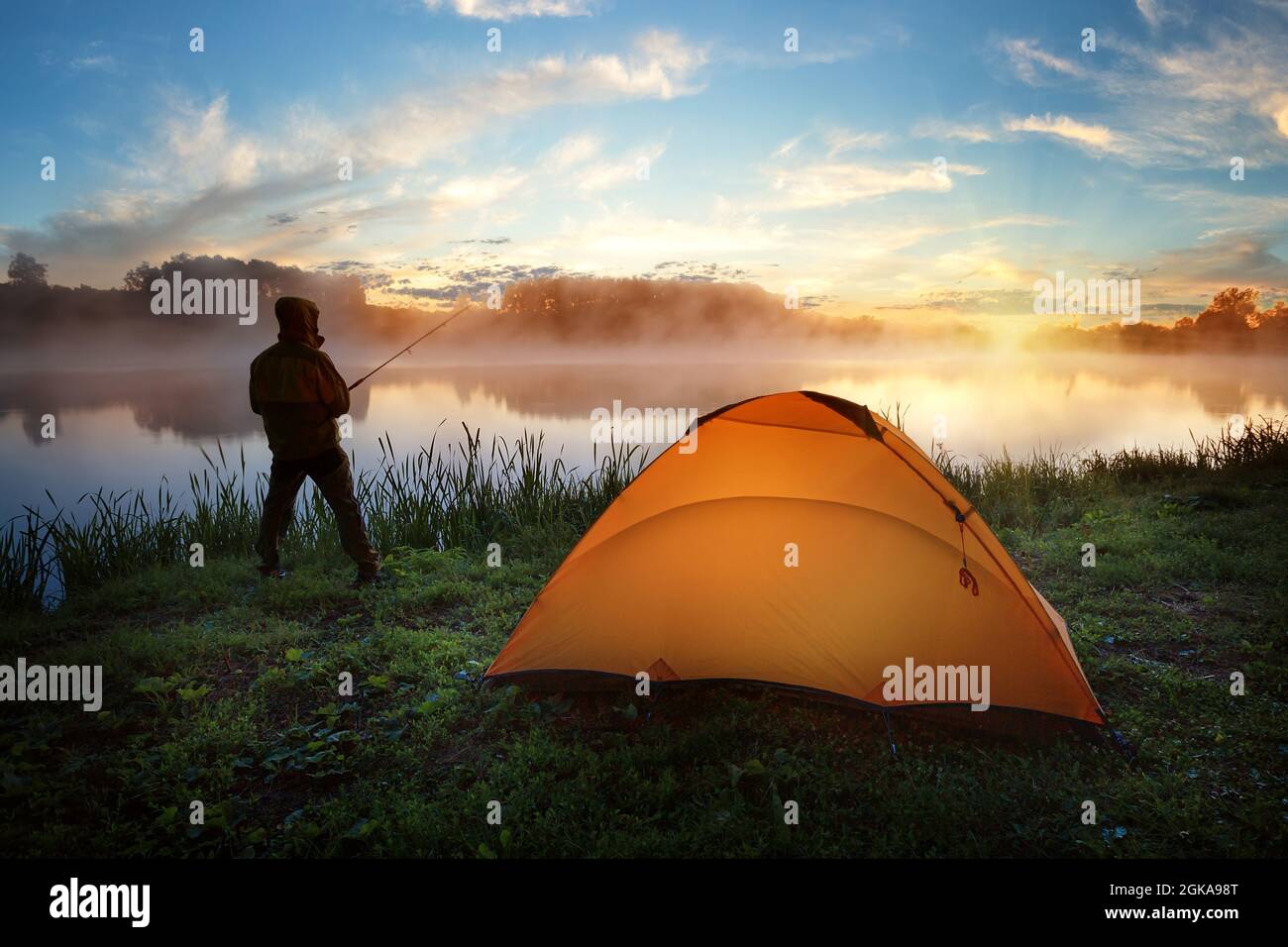 Fisherman near an orange tent on bank of misty river at sunset Stock Photo