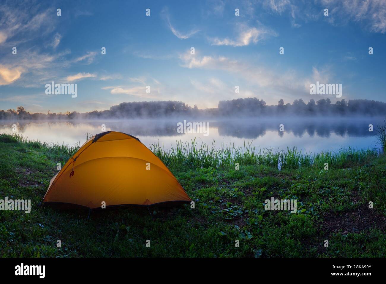 Tourist orange tent on green grass of foggy lake at sunrise Stock Photo