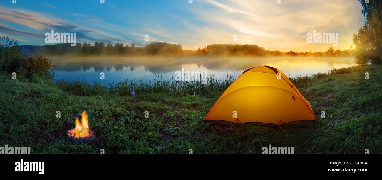 Orange tent with bonfire on bank of misty river at sunset Stock Photo