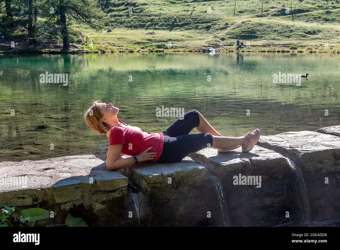 Blond woman lying in the sun on a low wall from which the water of Lago Blu flows, Aosta Valley, Italy Stock Photo