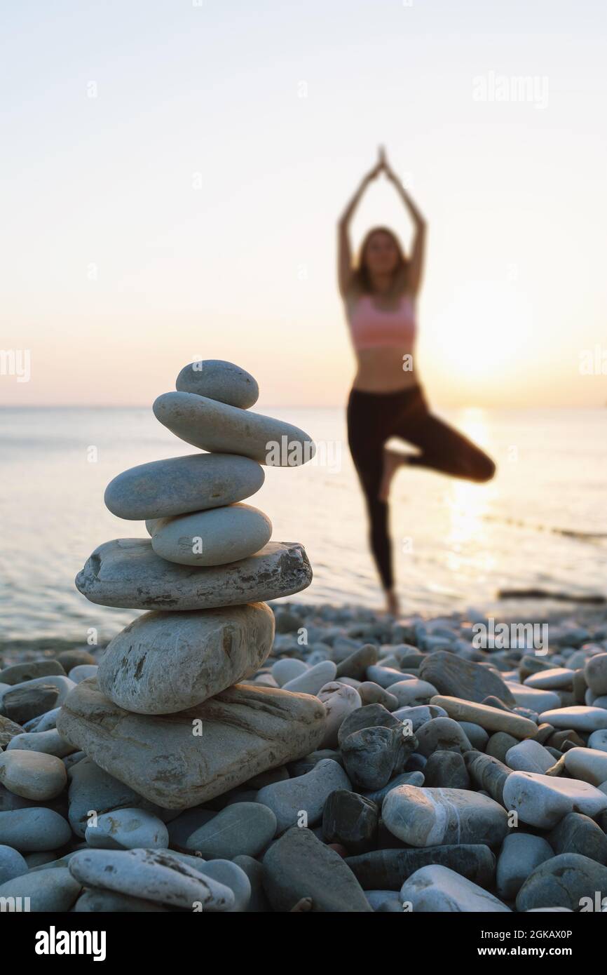 Balancing cairn of stones on the sea beach against the background of a woman out of focus performing the vrikshasana exercise, tree pose Stock Photo