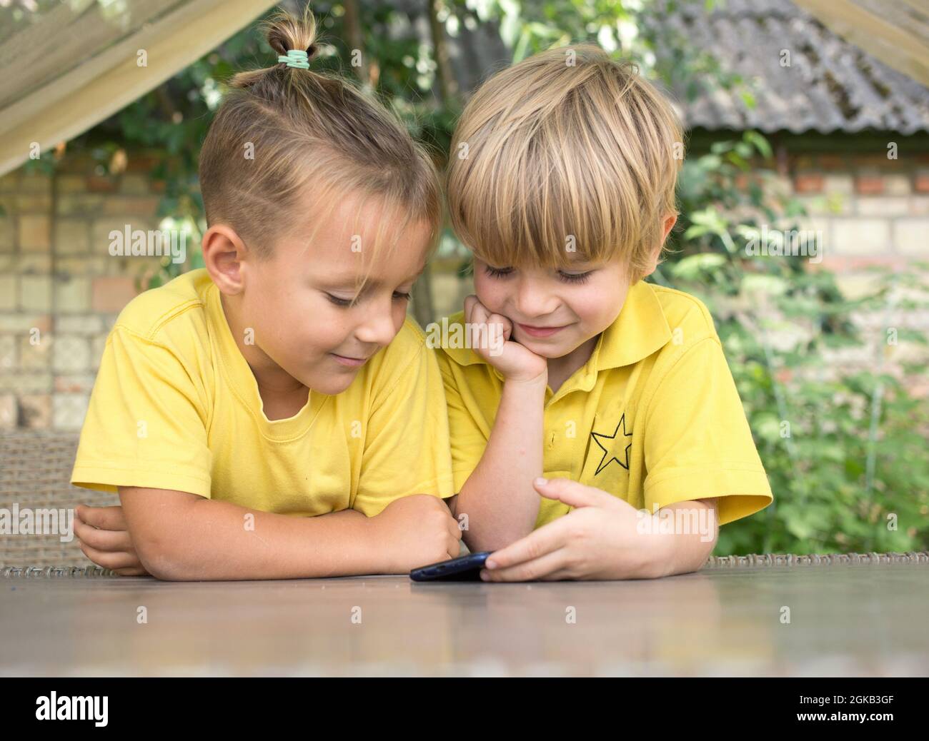 Cute 5 year old boys in yellow T-shirts are looking at phone screen in front of them with enthusiasm. modern children, digital era, curiosity, passion Stock Photo