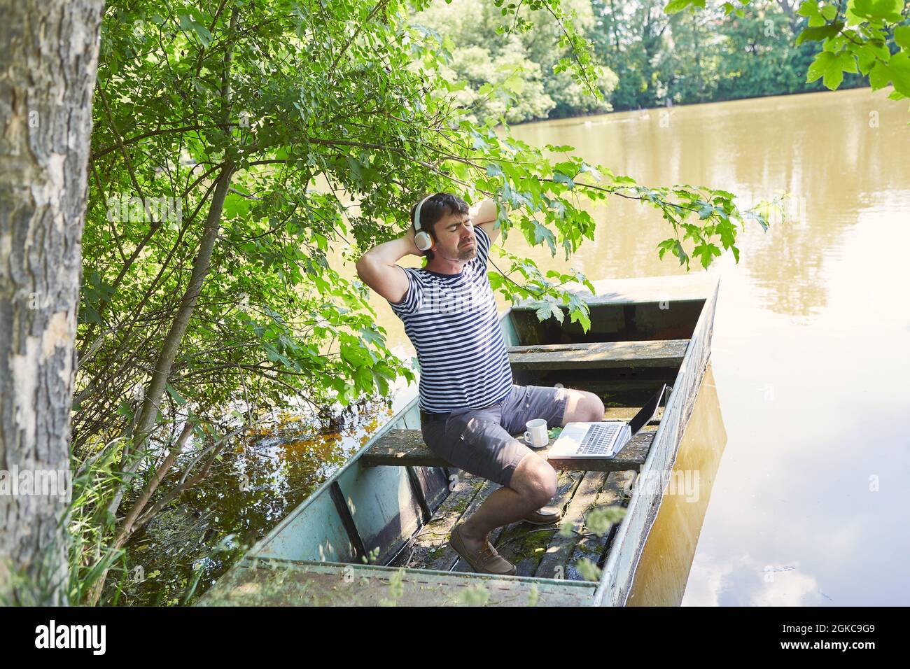 Freelancer with laptop PC sits relaxed on an old boat at the lake in nature Stock Photo