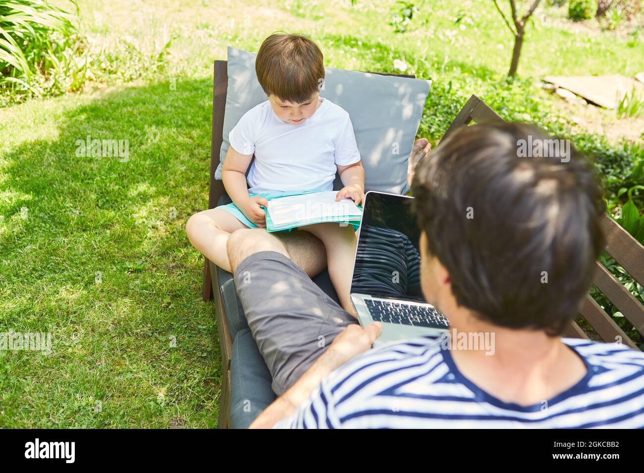 Father works on laptop computer online in the garden in summer with son doing it Stock Photo