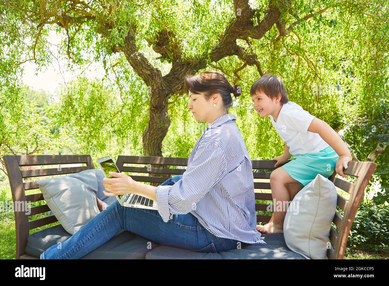 Single mother works on laptop PC in the garden with curious son Stock Photo
