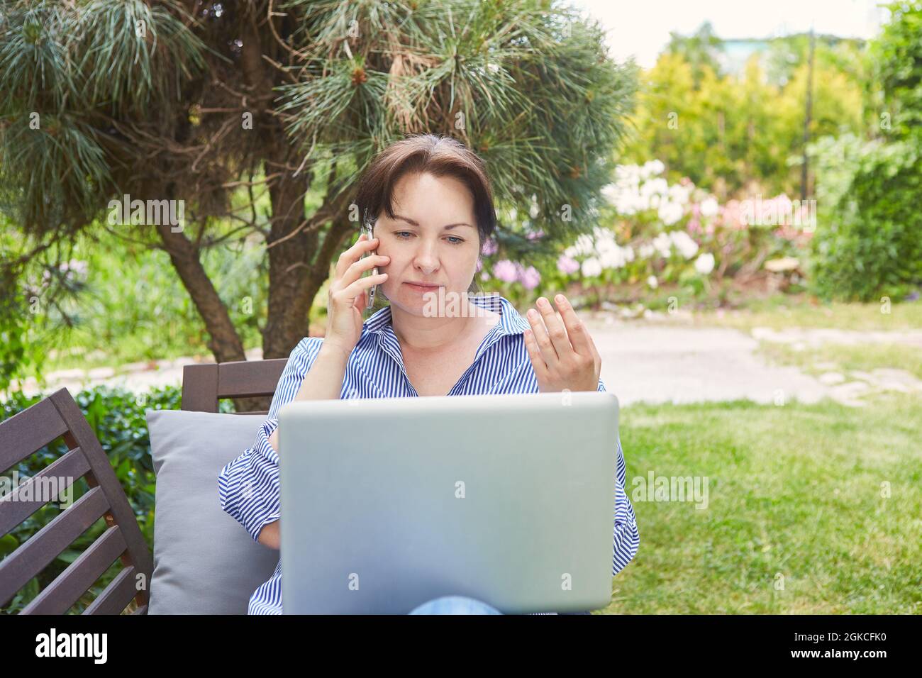 Business woman on laptop computer in green garden in summer making mobile phone call Stock Photo