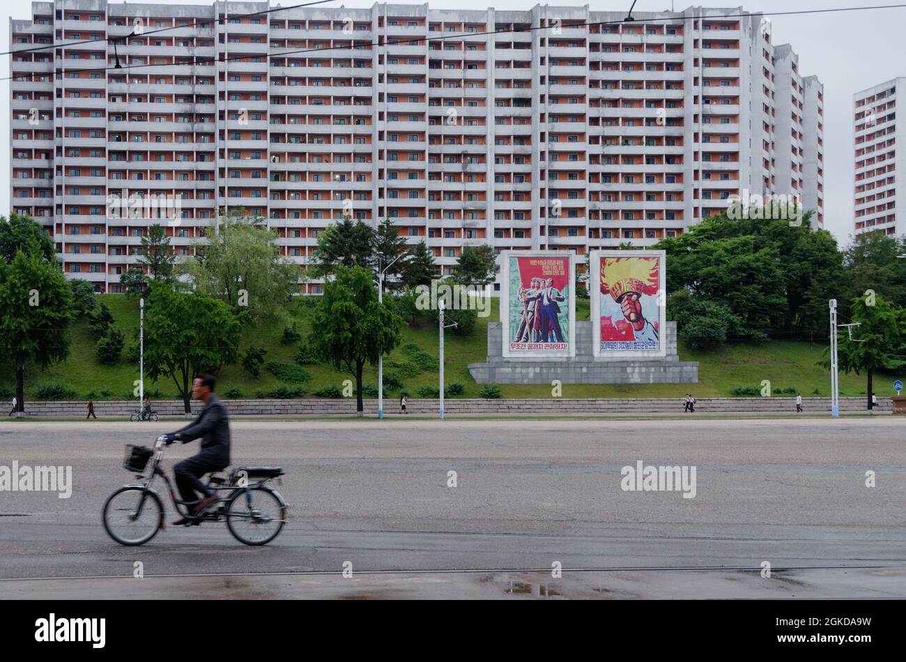Street photography in Pyongyang, a cyclist with propaganda posters and a housing block behind him. A wide road, no cars. Stock Photo