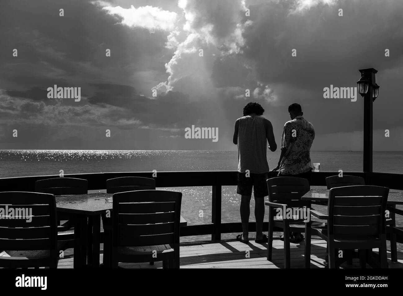 Two friends standing on a wooden terrace, enjoying the view of the sea, after a rainy episode. The sun pierces the clouds. Stock Photo