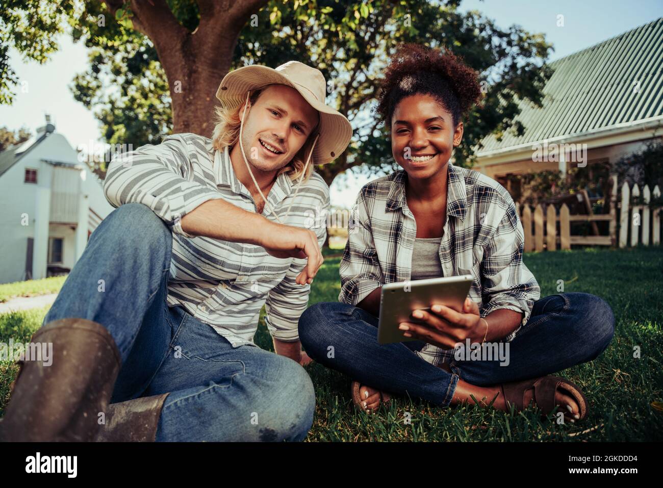 Mixed race couple smiling while enjoying time together sitting on grass scrolling through images on digital device  Stock Photo