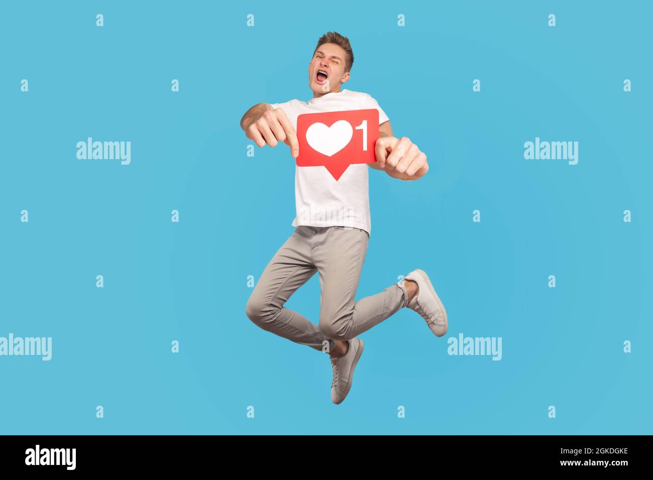 Happy successful overjoyed young man holding red social media like icon, jumping with happiness, screaming yes, I did it, rejoicing winning. indoor st Stock Photo