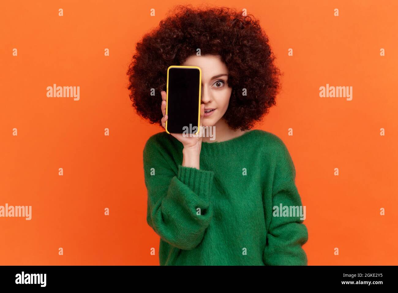 Attractive young adult woman with Afro hairstyle wearing green casual style sweater covering eye with cell phone with empty display. Indoor studio sho Stock Photo