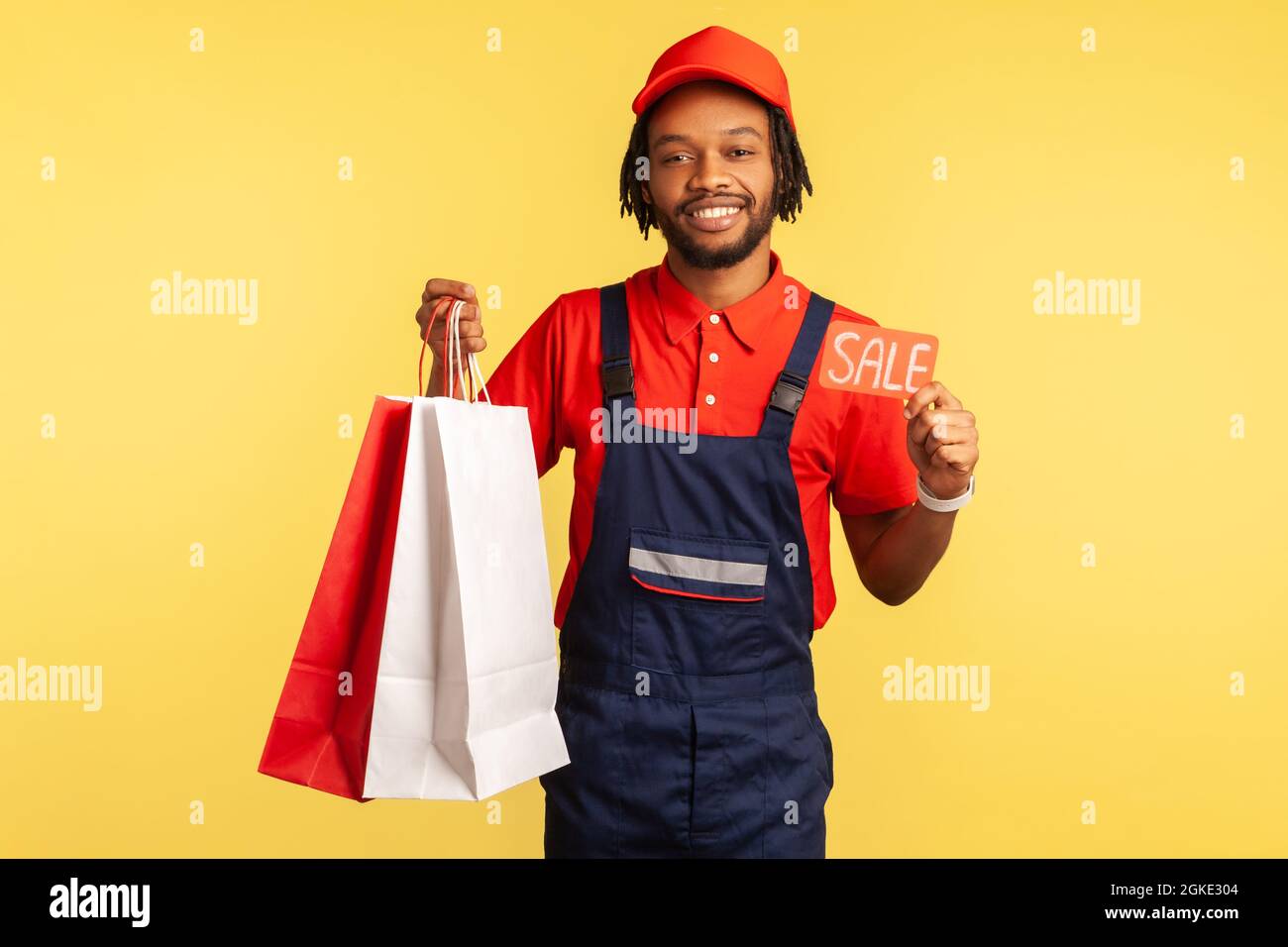 Smiling courier wearing uniform holding shopping bags from fashion store and showing sale coupon, sales at online orders and delivery. Indoor studio s Stock Photo
