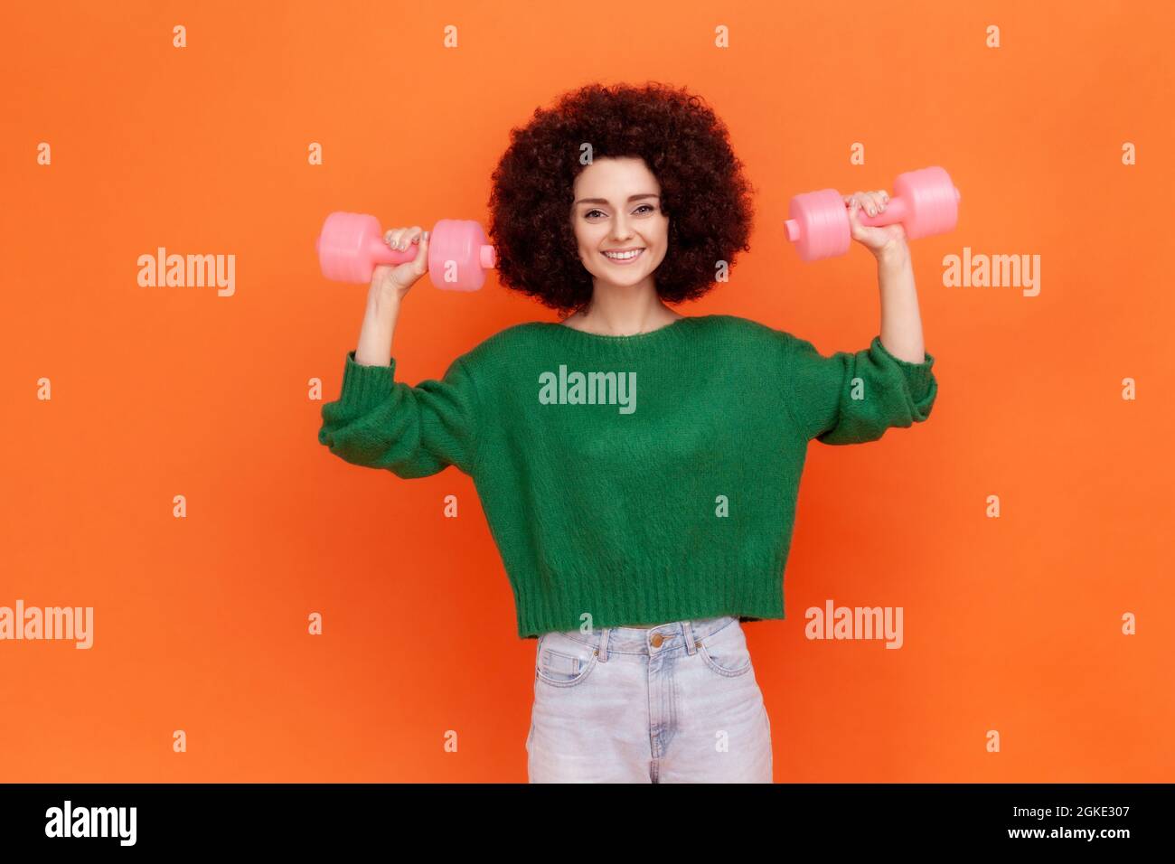 Positive woman with Afro hairstyle wearing green casual style sweater raising arms with pink dumbbells, training biceps and triceps, healthy lifestyle Stock Photo
