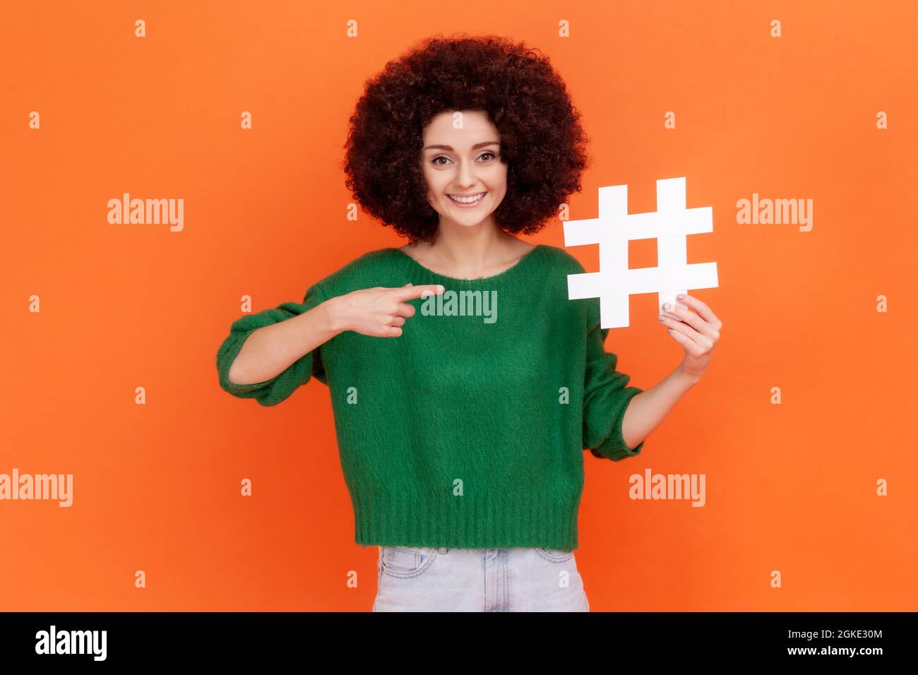 Viral hashtag and successful blogging. Smiling woman with Afro hairstyle wearing green casual style sweater pointing at large white hash symbol. Indoo Stock Photo