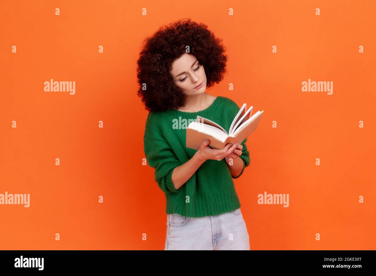 Attractive concentrated woman with Afro hairstyle wearing green casual style sweater reading interesting book, being very attentive. Indoor studio sho Stock Photo