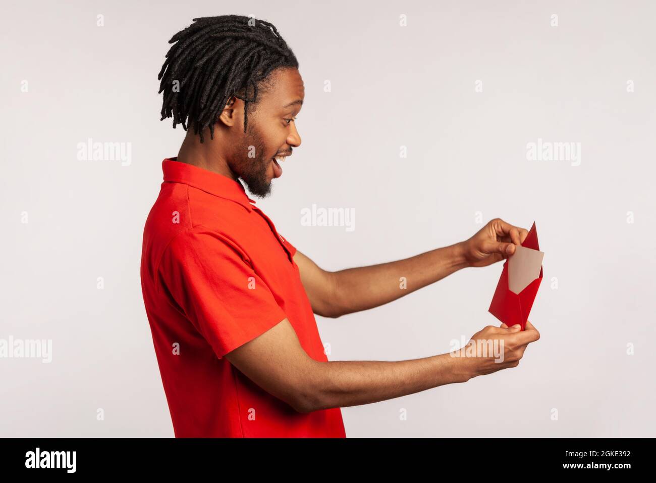 Side view of positive man with dreadlocks wearing red casual style T-shirt, opening letter in red envelope, holding greeting card and feels happy. Ind Stock Photo