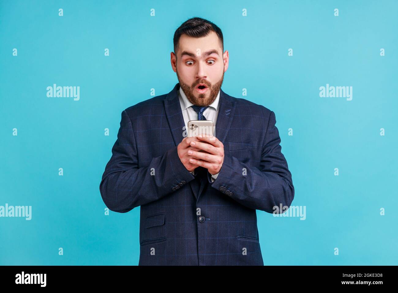 Bearded man wearing official style suit expressing shock while using cell phone, online mobile application, searching web, astonishing news. Indoor st Stock Photo