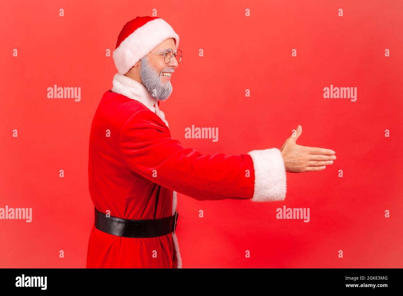 Side view of positive elderly man with gray beard wearing santa claus costume stretching his hand welcoming guests to new year party, hospitality. Ind Stock Photo