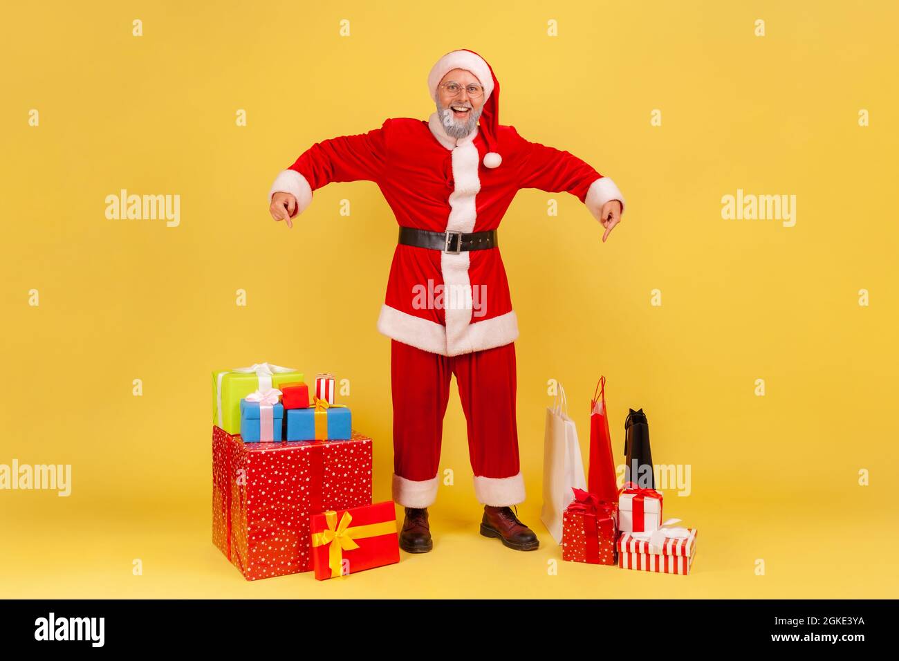 Full length portrait of smiling elderly man with gray beard wearing santa claus costume pointing at many present boxes for Christmas holidays. Indoor Stock Photo