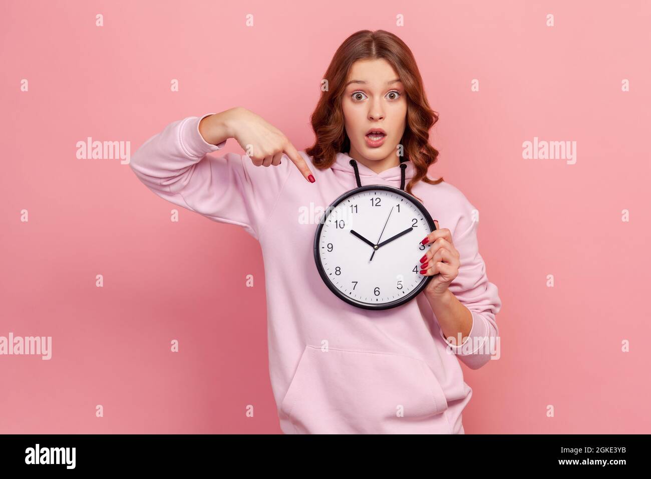 Portrait of shocked curly haired teen girl in hoodie pointing big clock and looking displeased irritated, showing wall watches to hurry up. Indoor stu Stock Photo