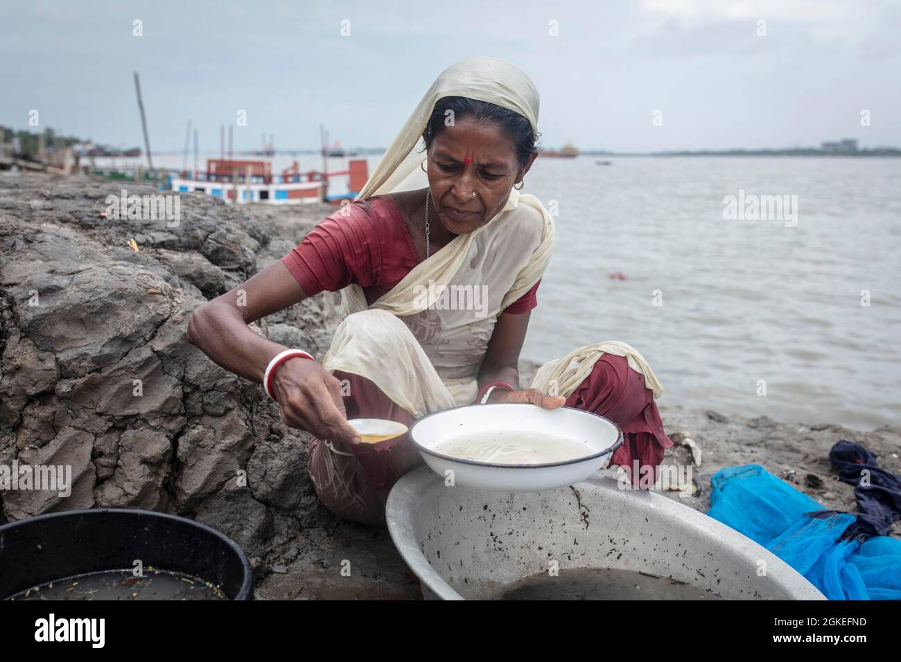 A woman sieves river water from wash bowls on a muddy embankment, shrimp larvae remain in the sieve to be sold to a shrimp farm, Mongla, Sundarbans Stock Photo
