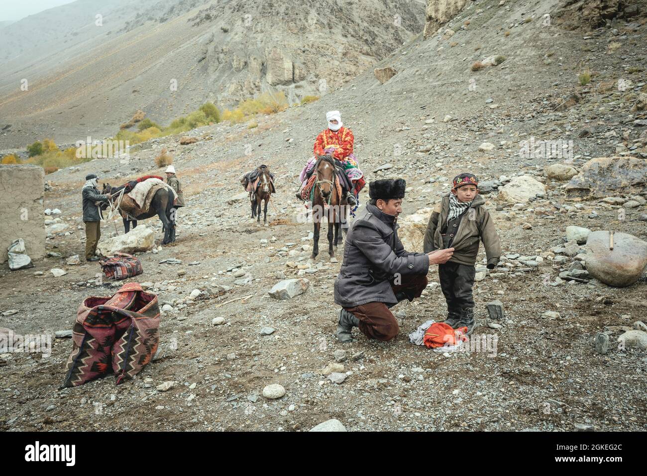A man puts on a jacket for his son, a horse is loaded in the background, a woman sits on a horse, Kyrgyz nomads on their way to the hospital of Stock Photo