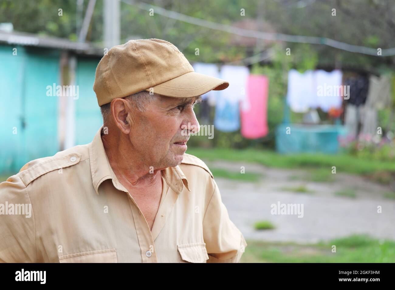 Elderly man standing in rural yard on background of well and hanged laundry. Concept of life in village, old age Stock Photo