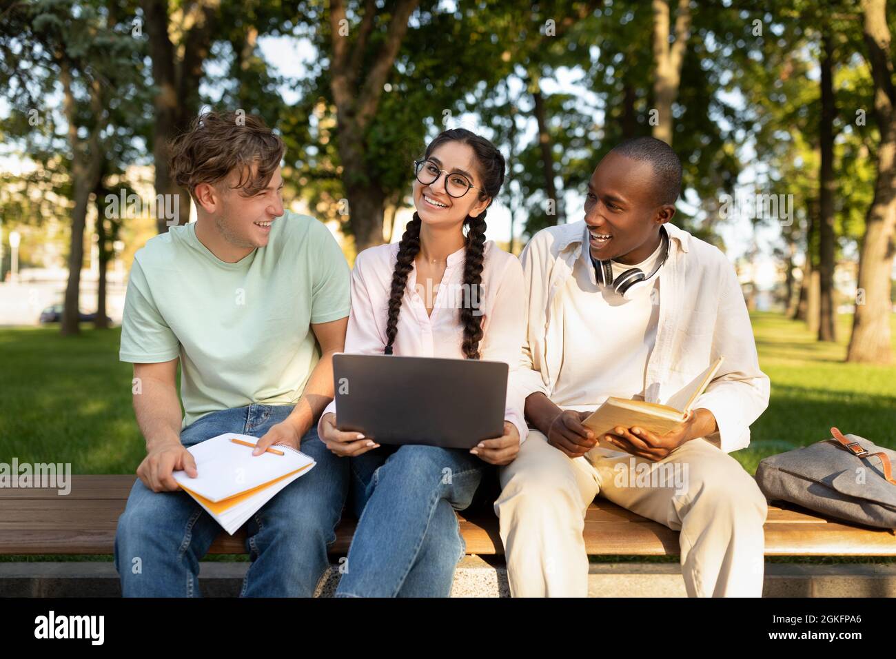 Diverse students friends learning sitting on bench, using laptop outdoors in university campus Stock Photo