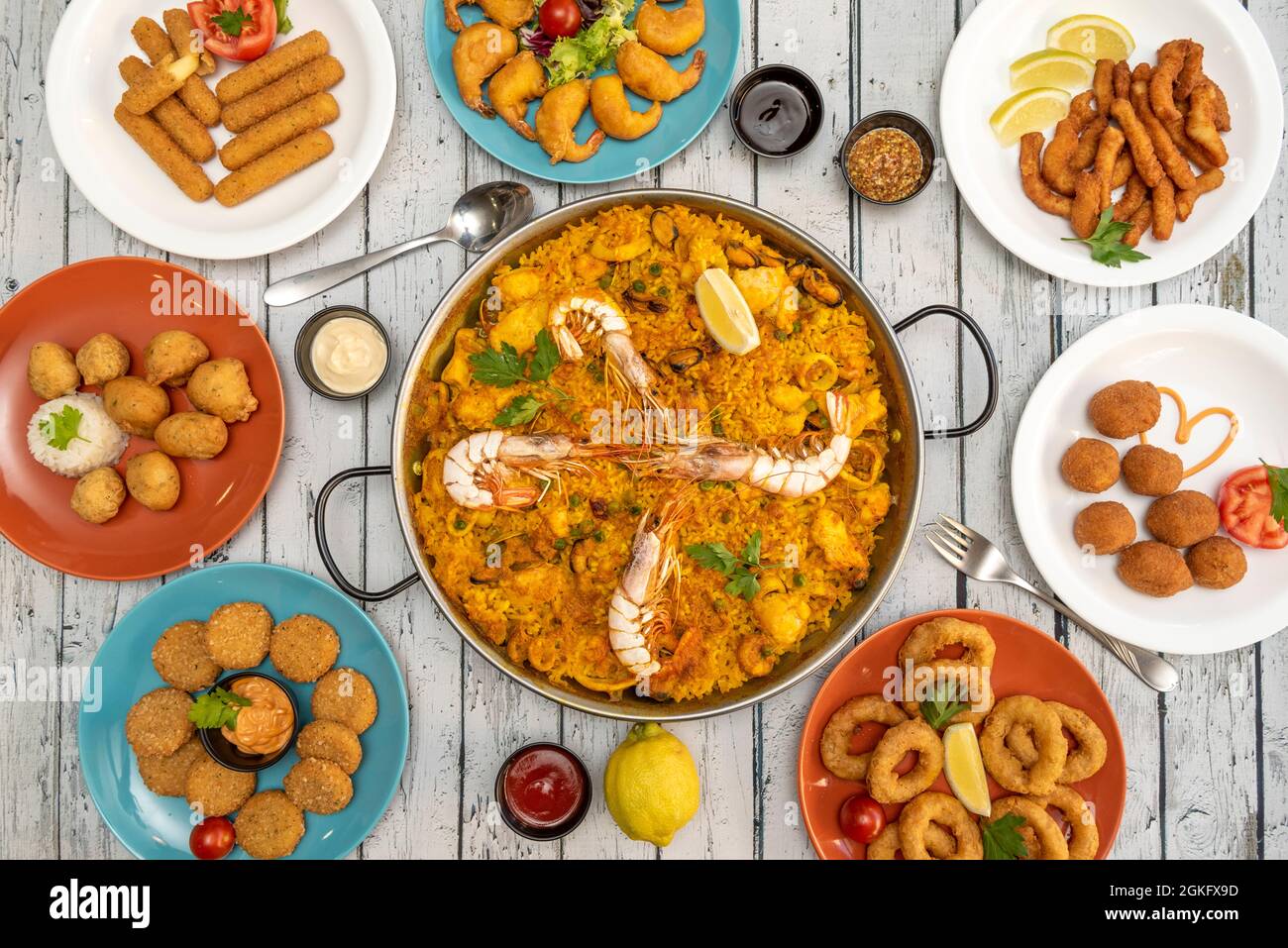 Top view image of appetizer plates and a seafood paella in the center on a wooden table with cutlery and sauces Stock Photo