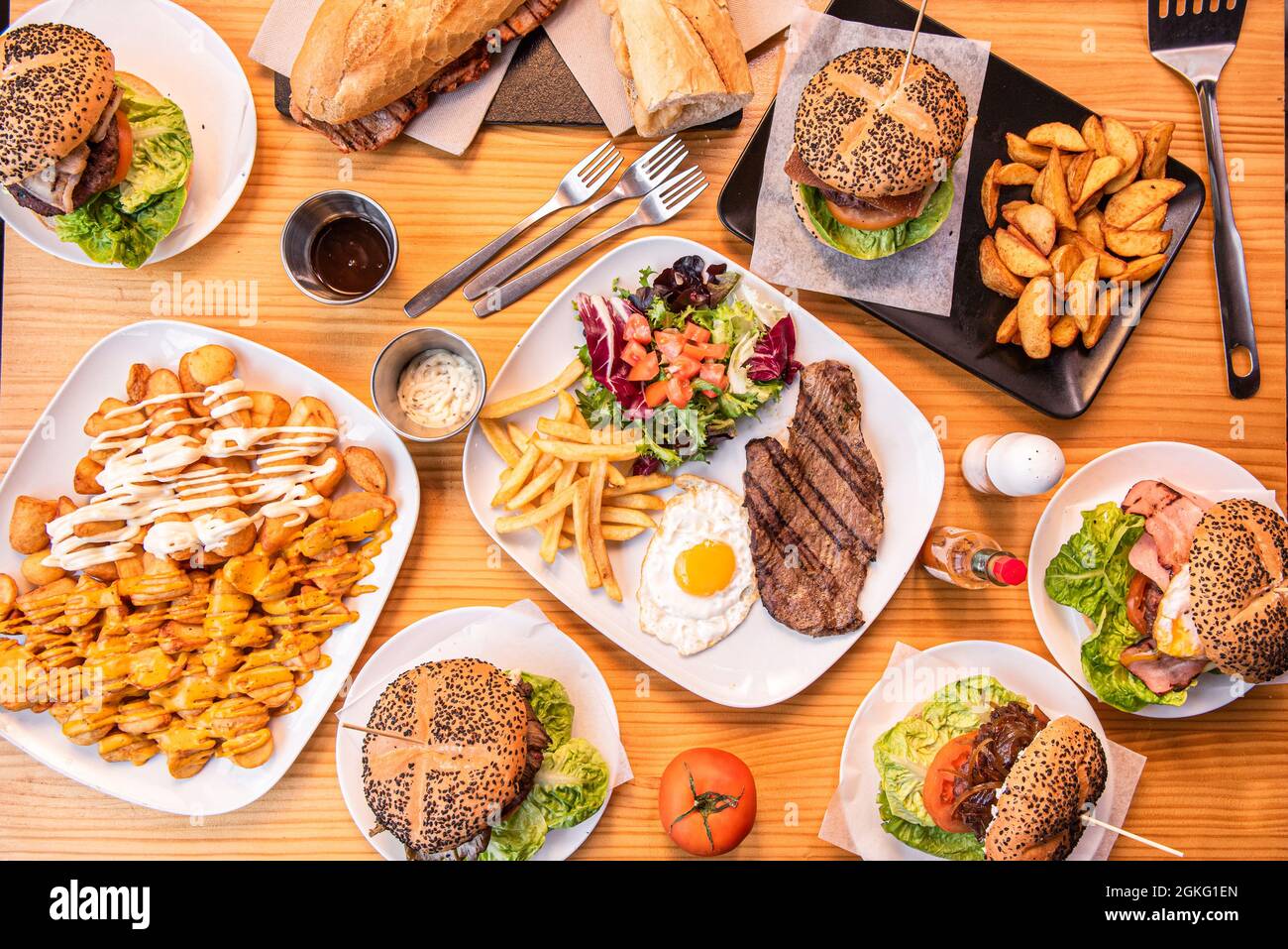 Set of popular Spanish dishes and hamburgers on wooden table. Loin and cheese sandwich, steak and egg, patatas bravas, fresh tomatoes, forks, deluxe p Stock Photo