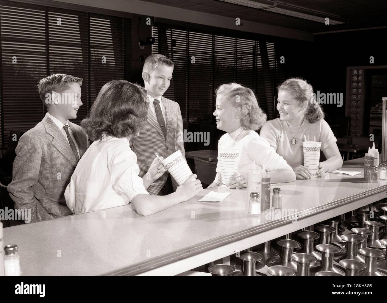 1950s TWO BOYS AND THREE GIRLS TEENAGERS TOGETHER AT SODA FOUNTAIN COUNTER TALKING FLIRTING DRINKING MILK SHAKES - f4510 HAR001 HARS FOUNTAIN RELATIONSHIP SOCIAL OLD TIME FLIRT NOSTALGIA OLD FASHION T JUVENILE STYLE COMMUNICATION LAUGH PLEASED JOY LIFESTYLE FEMALES 5 COPY SPACE FRIENDSHIP HALF-LENGTH PERSONS MALES TEENAGE BOY CONFIDENCE B&W DATING SUIT AND TIE HAPPINESS CHEERFUL LEISURE AND EXCITEMENT AT ATTRACTION SMILES COURTSHIP JOYFUL STYLISH TEENAGED POSSIBILITY JUVENILES PRE-TEEN SHAKES SOCIAL ACTIVITY TOGETHERNESS BLACK AND WHITE CAUCASIAN ETHNICITY COURTING HAR001 OLD FASHIONED Stock Photo