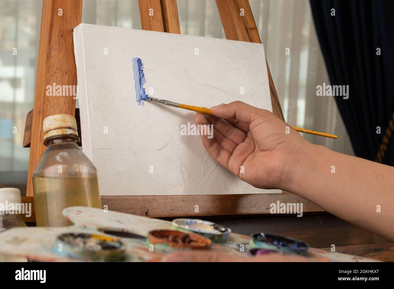 hand of a person holding a brush while painting on a wooden canvas and holding a palette, art elements in work area with natural day light, creative Stock Photo