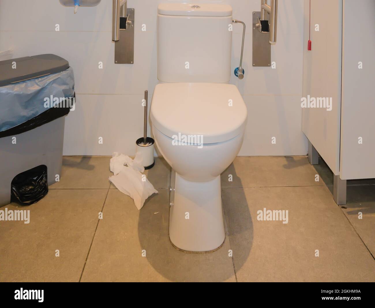View into a public toilet, lid closed, armrests folded up against the wall. On the left of the picture there is a large trash can with garbage bags. Stock Photo