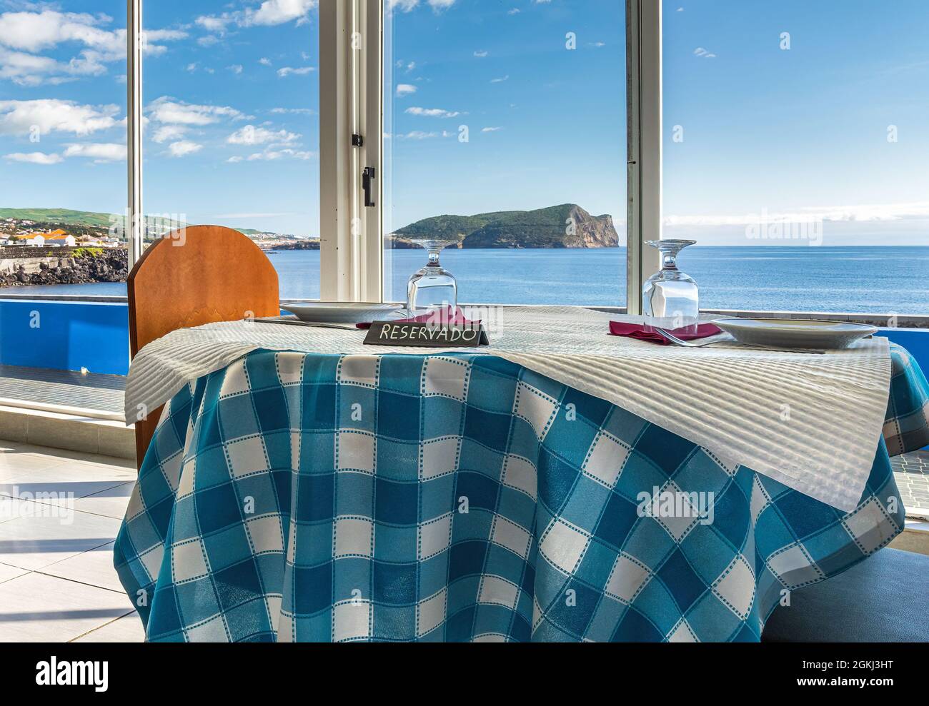 Reserved table in a restaurant with a view of the coast of Terceira, Azores, with a blue table cloth and a sign that says “reservado.” Stock Photo