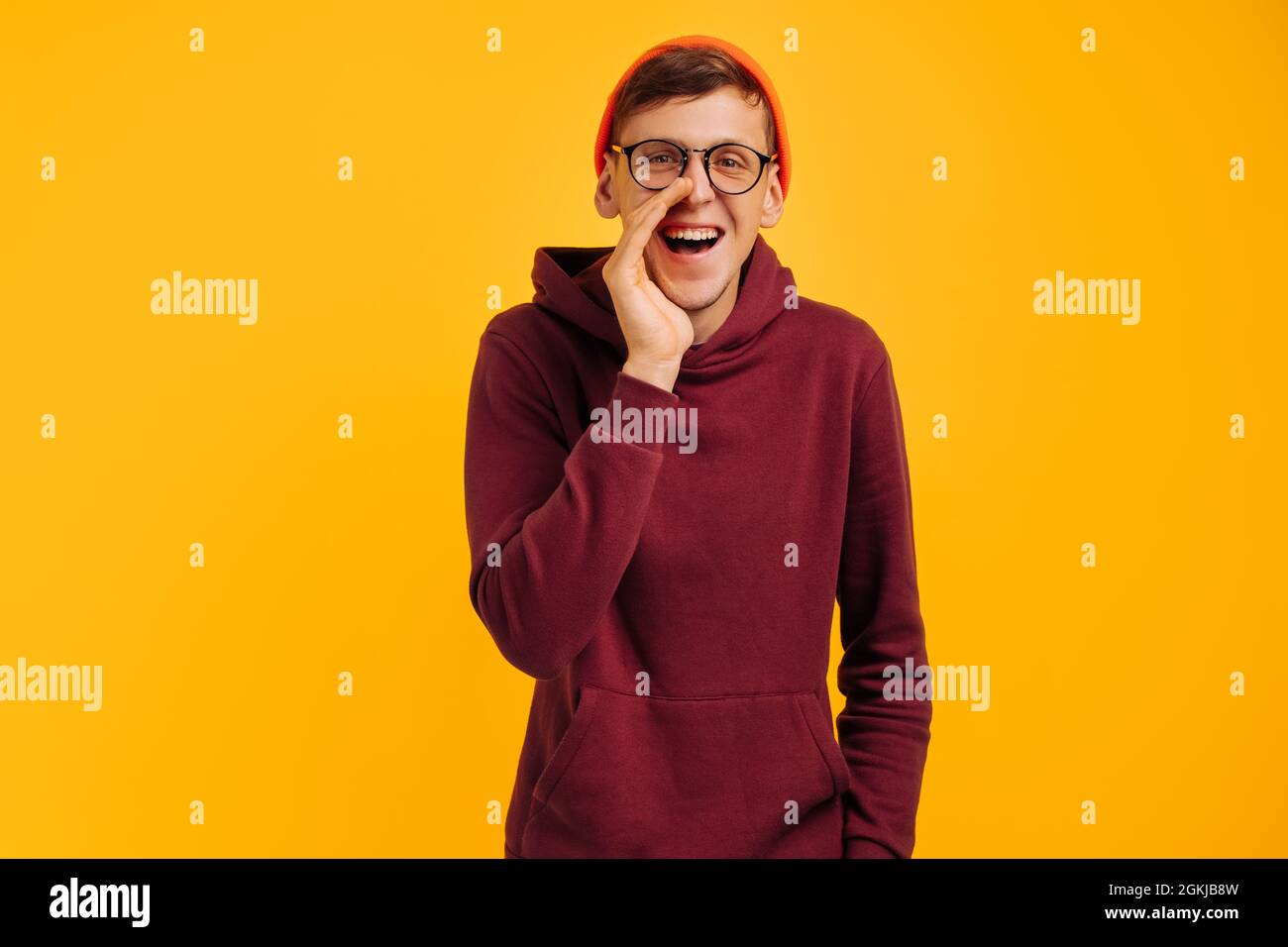 handsome man says something in secret, a guy in an orange hat and a red sweater is gossiping, talking in gray, on a yellow background Stock Photo