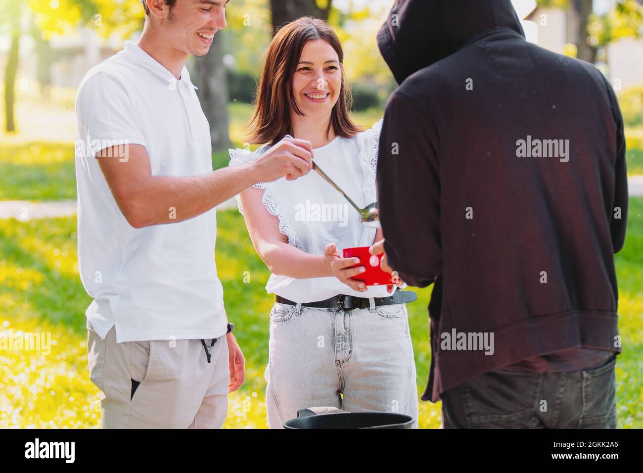 Happy couple making volunteer work of feeding homeless man in a hoodie with  a hood on his head, giving to him a big red bowl with some soup Stock Photo  - Alamy