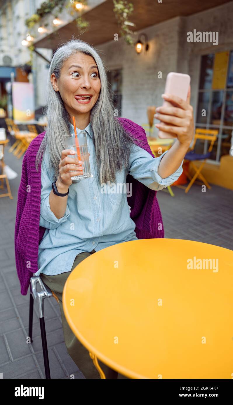 Funny senior Asian woman takes selfie grimacing on outdoors cafe terrace Stock Photo