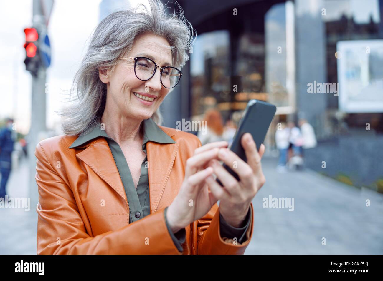 Positive mature lady with glasses uses phone standing on city street Stock Photo