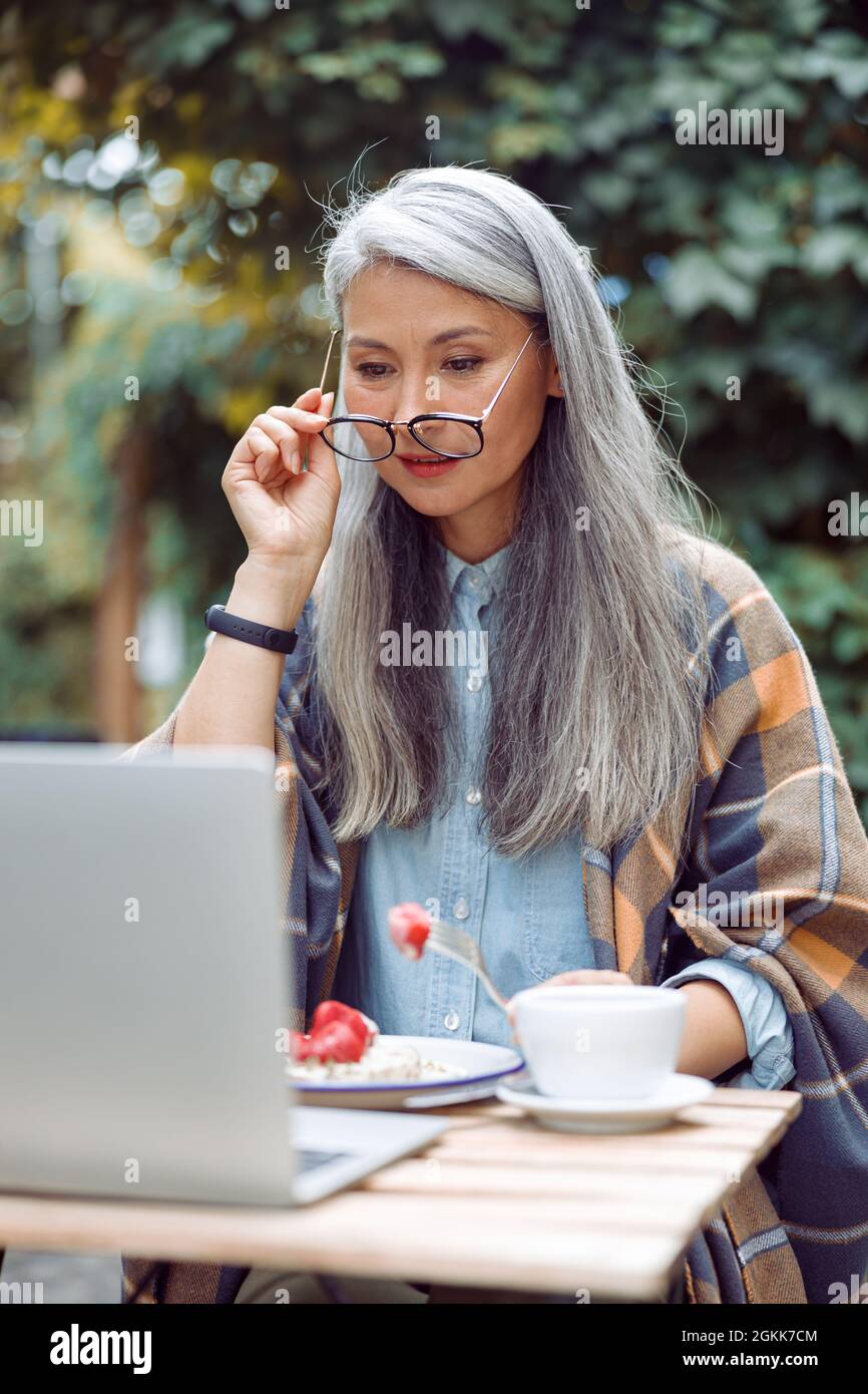 Concentrated mature Asian lady with glasses and strawberry looks at laptop at table outdoors Stock Photo