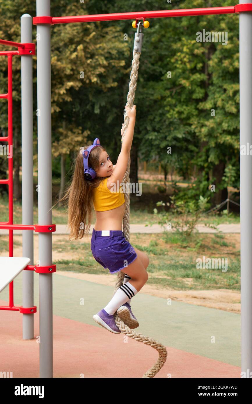 Sporty little girl workout outdoor on sports ground. Schoolgirl preteen  wearing fashionable sportswear and wireless headphones listening to music  with pleasure enjoy activity climb rope Stock Photo - Alamy