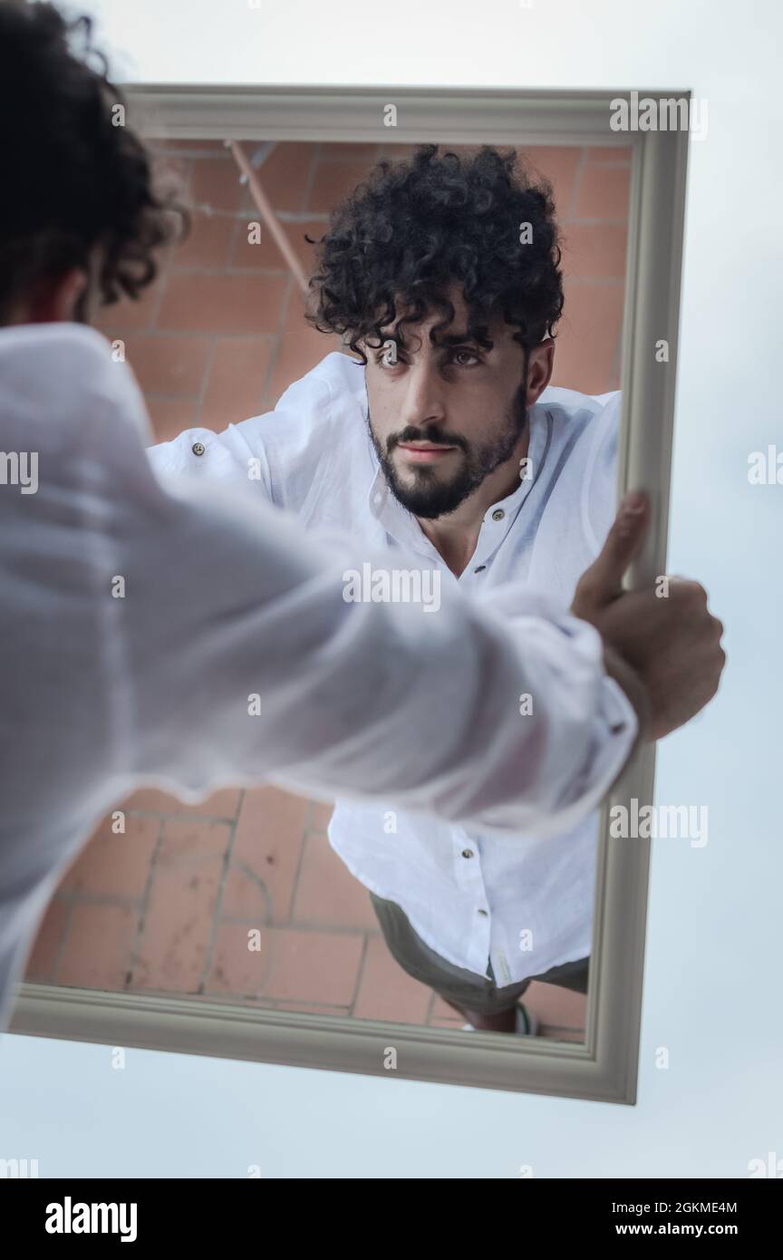portrait of a young man with beard and curly hair, who looks at himself  while holding a mirror in his hands Stock Photo - Alamy